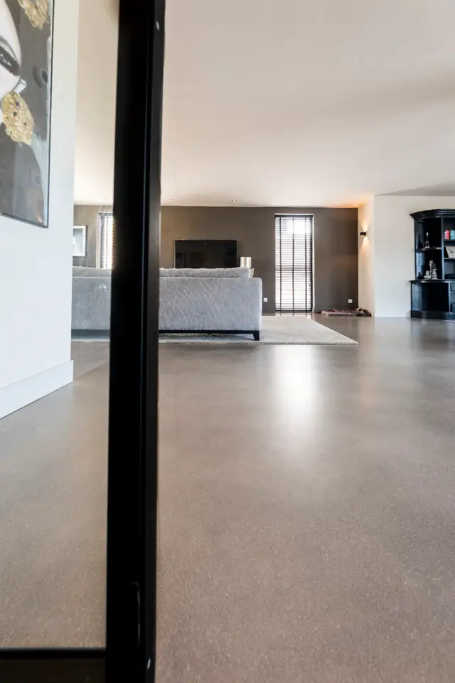 Spacious open-plan living area met een doorlopende naadloze vloer in warm grijs. Strakke interieurafwerking met minimalistisch design, natuurlijk licht en elegante meubelbezetting.