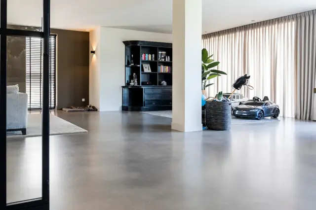 Strakke, open leefruimte met een grote naadloze vloer in warme grijstint. Het interieur combineert minimalistisch design met luxe details: zwarte inbouwkast, marmeren accenten en veel natuurlijk licht door grote ramen.