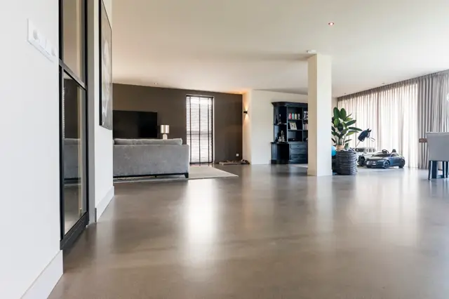 Ruime, licht inval woonkamer met grijze naadloze vloer die de hele ruimte visueel verbindt. Strakke architectuur met wit plafond, donkere accentwand en minimalistisch interieur.
