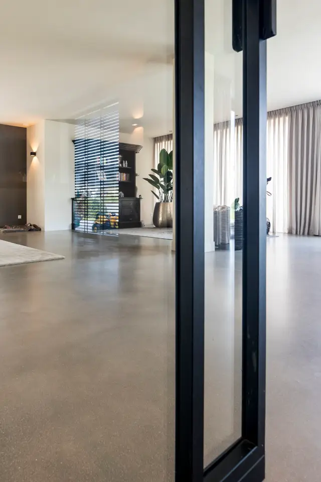 Groot, licht wooninterieur met doorlopende naadloze vloer in warm grijs. Strakke architectuur met zwarte stalen kolommen, minimalistische inrichting en natuurlijk licht door grote ramen met gordijnen.