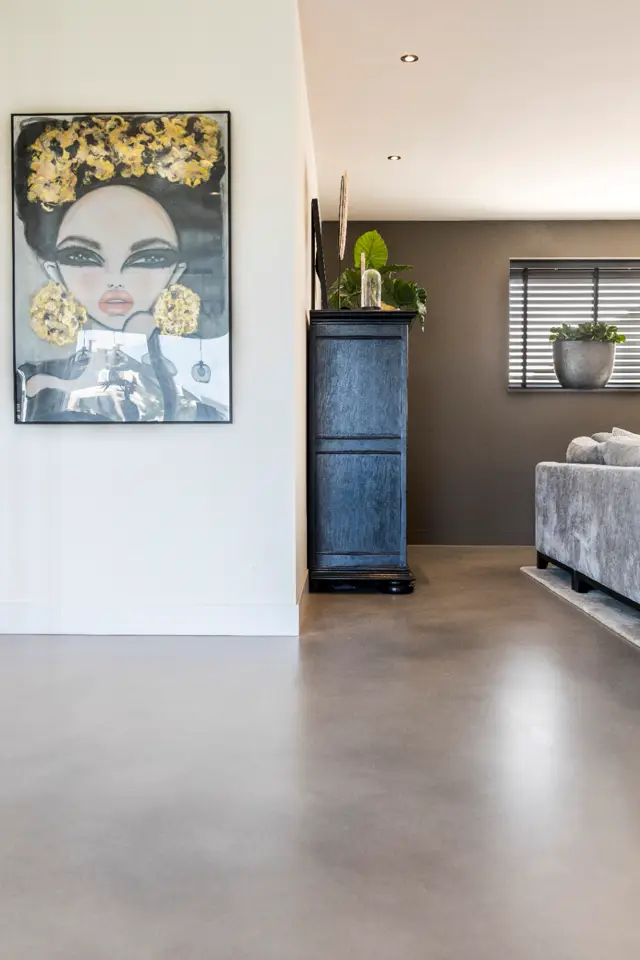 Ruime, lichte hal met naadloze vloer in warm grijs-beige. Minimalistische inrichting met kunstwerk aan witte wand, blauwe vintage kast en grijze stoelgroep.