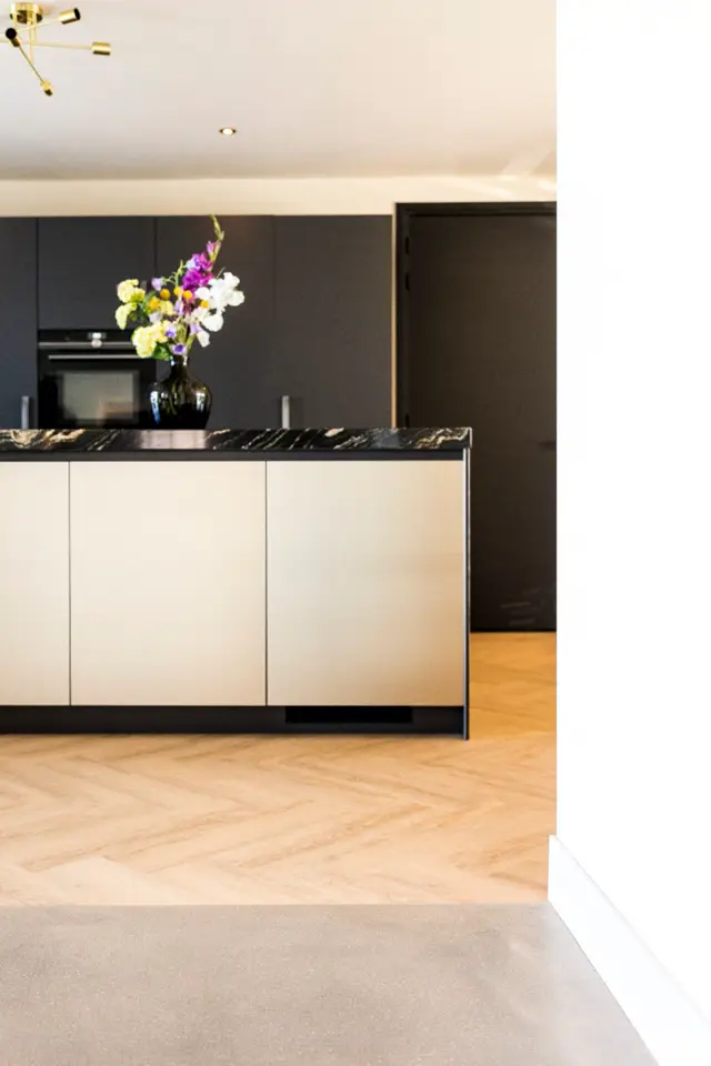 Strakke, moderne keukenopstelling met contrasterende zwarte wandafwerking en crèmekleurige kasten. Gouden designlamp en fris bloemstuk creëren elegante accenten boven de naadloze werkvlak.