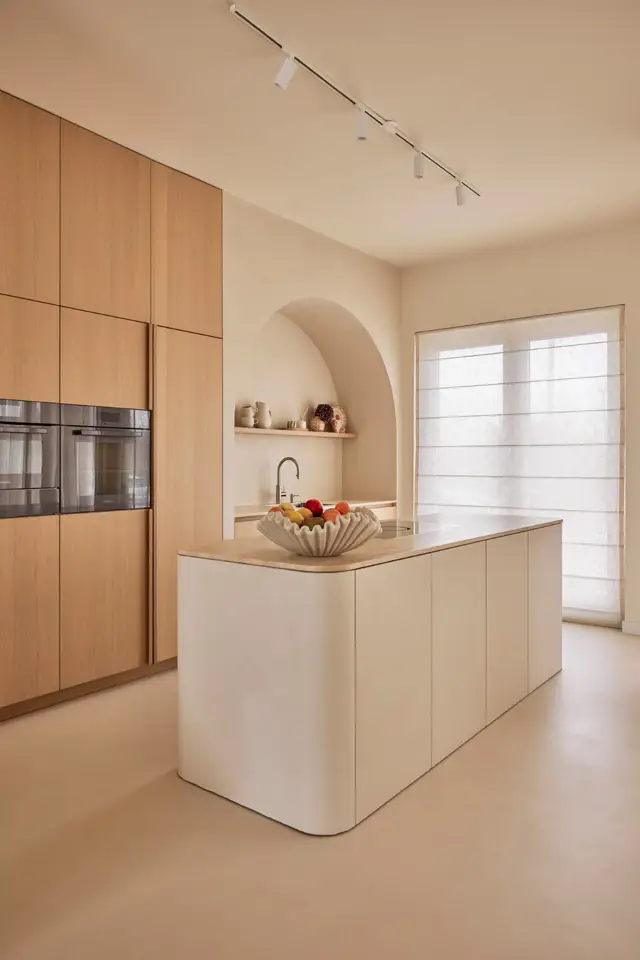 Serene keukenruimte met naadloze wand- en vloerwerk in warme beige tinten. Een sculpturaal eiland met afgeronde hoeken domineert de ruimte, geaccentueerd door een organische fruitschaal en sierlijke kraanwerk.
