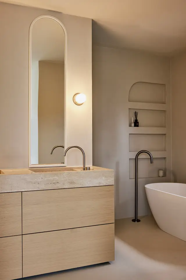 Een serene badkamer met naadloze wanden in warm beige, een elegant langwerpig spiegel met subtiele verlichting, houten wastafelmeubel met stenen blad, en ingegraven wandnissen. De ruimte ademt rust en verfijning uit met scherpe lijnen en perfecte proporties.