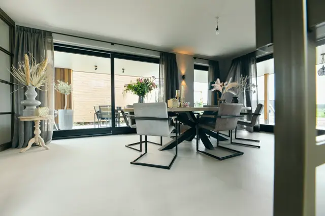 Ruime, moderne eetkamer met een strakke naadloze vloer in licht grijs. De ruimte ademt luxe en rust uit dankzij grote raampartijen, minimalistisch meubilair en verfijnde decoratie.