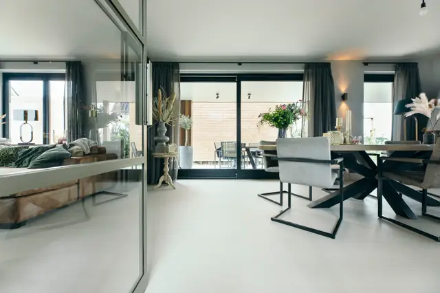 Ruim, licht interieur met naadloze vloer in neutrale tint die zich naadloos uitstrekt van het woongedeelte naar slaapkamer en terras. Strakke lijnen, minimale dekking en grote raampartijen creëren een serene, opgeruimde uitstraling.