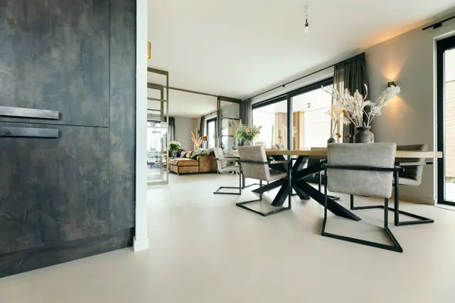 Ruime, heldere woonkamer met een vrijdragende tafel en grijze stoelen. De naadloze vloer loopt naadloos door naar de woonzone, terwijl een donkere naadloze wand links contrast biedt. Natuurlijk licht stroomt in via grote raampartijen.