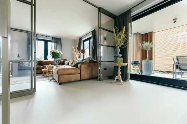 Ruime, lichte woonkamer met naadloze vloer in warm wit, grote raampartijen met donkere framewerken en minimalistisch ingericht met bruin lederen meubels. De ruimte oogt schoon, gestructureerd en helder.