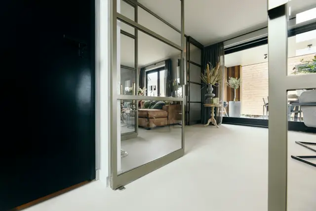 Ruime, lichte hal met naadloze vloer en grote glazen schuifdeuren die uitkijken op een modern woonvertrek. De ruimte ademt elegantie en openheid met strakke lijnen en minimalistische detaillering.