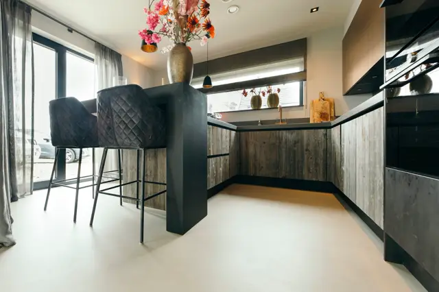 Strakke, open keuken met houtstructuur kasten, zwart eiland en lichte naadloze vloer. Grote ramen zorgen voor natuurlijk licht en een luchtig gevoel.