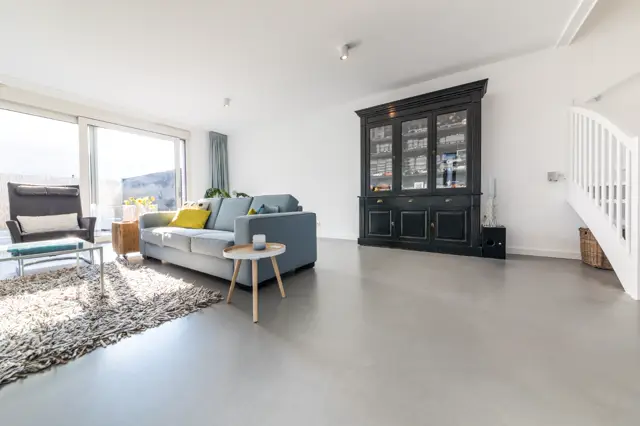 Ruim, minimalistisch ingerichte woonkamer met een naadloze lichte vloer die het interieur airy en schoon doet uitstralen. Natuurlijk licht, strakke lijnen en hoogwaardige afwerking.