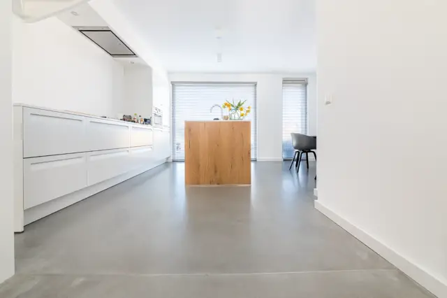 Strakke, witte keuken met houten keukeneiland op een groot, naadloos grijs oppervlak. Maximale rust en ruimtelijkheid door schone lijnen en overvloedige natuurlijke lichtinval.