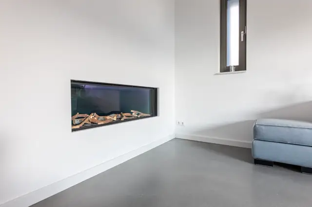 Strakke, lichte woonruimte met naadloze wand in wit, ingebouwde elektriache haard en grijze naadloze vloer. Het geheel ademt rust, openheid en hedendaagse luxe uit.
