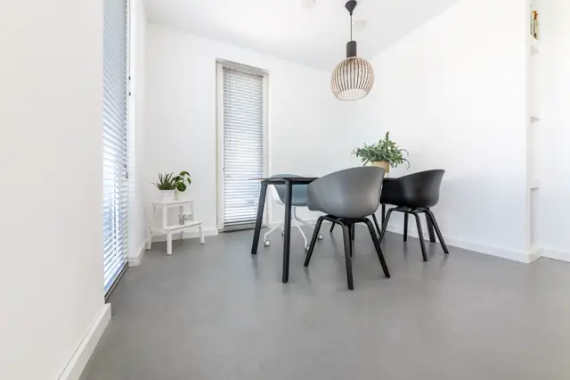 Schoon, licht eetgedeelte met strakke naadloze grijze vloer, witte wanden en zwart design meubilair. Het ruimtegevoel wordt versterkt door grote ramen en doordachte verlichting.