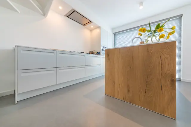 Strakke, lichte keuken met witte kastenwand en warm houten eiland. Naadloze vloer loopt ononderbroken door de ruimte, ondersteund door perfecte belichting en grote ramen met natuurlijk licht.