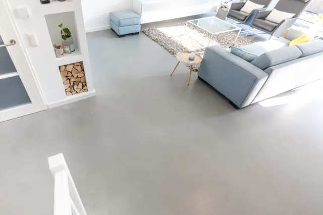 Ruime, lichte woonkamer met een naadloze grijze vloer die naadloos aansluit op een open leefruimte. Minimalistisch ingericht met blauwe accenten, natuurlijk licht en strakke lijnen.