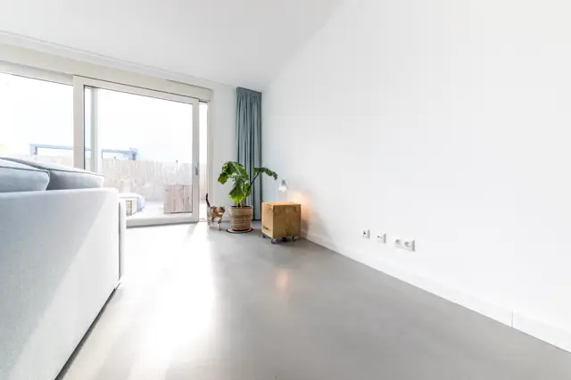 Ruime, minimalistische woonkamer met grote raampartijen en een strakke naadloze vloer in licht grijs. De ruimte ademt rust en luxe uit dankzij het hoge plafond, witte wanden en doordachte styling.