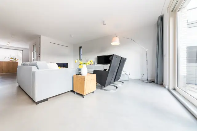 Ruim, minimalistisch ingerichte woonkamer met grote raampartijen, naadloze lichte vloer en wanden. Het interieur ademt rust uit met wit en natuurlijke houttinten.