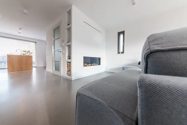 Ruim, licht interieur met glanzende naadloze vloer in warm grijs. Strakke witte wanden, geïntegreerde haard en hoogwaardige afwerking creëren een serene, geordende luxe-uitstraling.