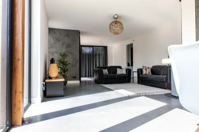 Ruime, lichte woonkamer met strakke naadloze vloer in warm grijs. Natuurlijk daglicht creëert scherpe geometrische schaduwen; minimalistisch ingericht met zwarte meubels en natuurlijke accenten.