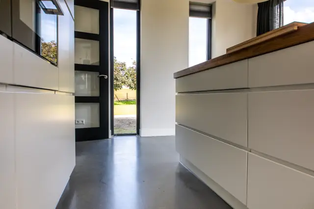 Serene hal met gladde donkergrijze naadloze vloer, witte wanden en subtiele inbouwkasten. Groot raamfront met zwart frame biedt uitzicht op tuinlandschap en zorgt voor optimaal natuurlijk licht.