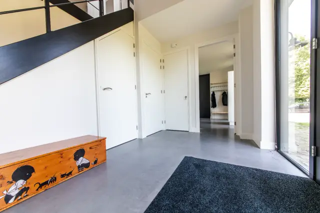 Ruime, lichte hal met gladde grijze naadloze vloer die zich naadloos uitstrekt naar de aangesloten ruimtes. Strakke witte kasten, natuurlicht van buiten en minimalistisch interieur creëren een opgeruimde, luxe uitstraling.