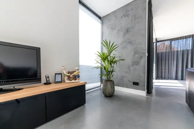 Strak ingerichte bedrijfsruimte met lichte natuurinval, grijze naadloze wand en vloer, minimaal meubilair en een enkele groene plant als designelement. De ruimte straalt rust en ordening uit.