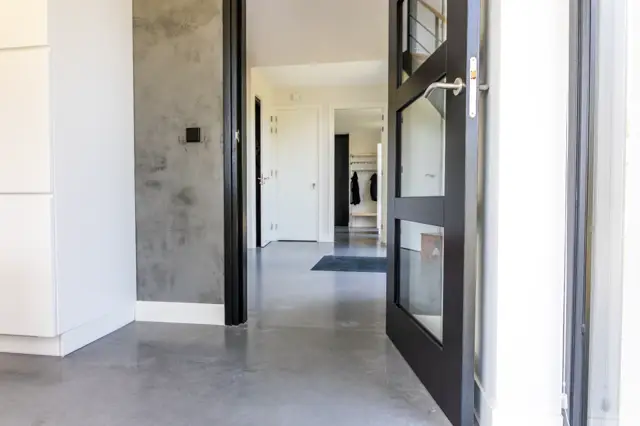 Strakke entreehal met grijze naadloze vloer, witte wanden en zwart-metallic deurkozijn. Clean design met minimalistische uitstraling en optimaal gebruik van licht.