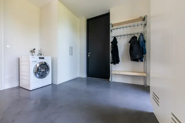 Schone, lichte wasruimte met een donkergrijze naadloze vloer en witte wanden. Minimalistische inrichting met een wasmachine, garderobe-element en organisch geordende ruimte.