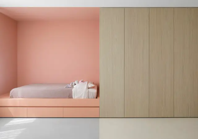 Minimalistisch slaapkamerinterieur met een geïntegreerd roze bed in nis, naadloze lichte vloer en natuurlijke houten kastenwand. Clean design met scherpe lijnen en perfecte proporties.