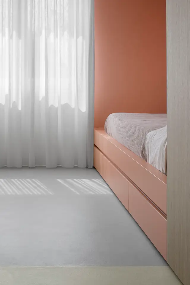 Serene slaapkamer met lichte naadloze vloer, grote ramen met diffuus daglicht, en een boldface terracotta-gekleurde wand die contrasterend tegen de rust van het interieur staat.