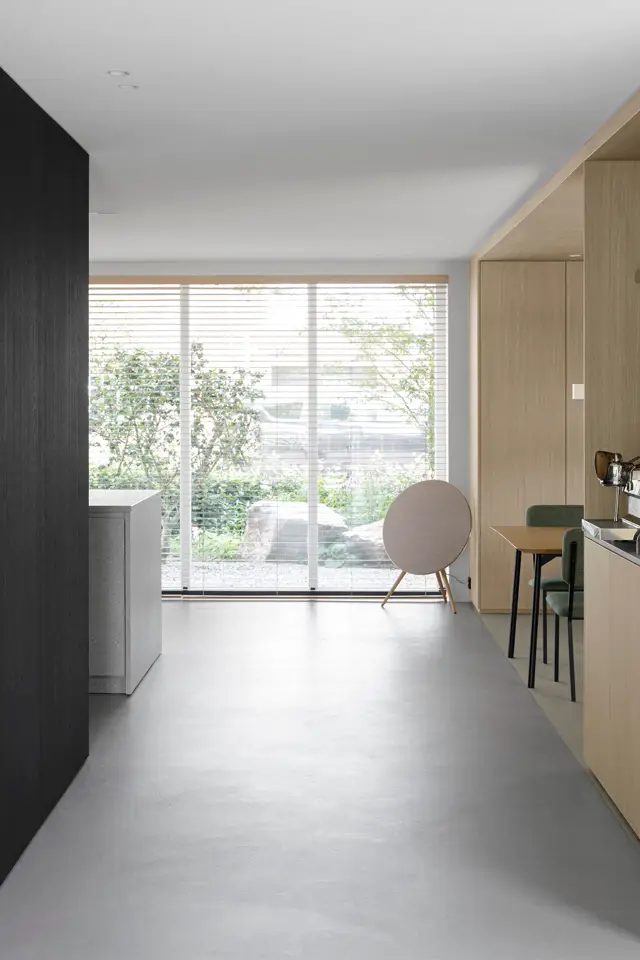 Licht en ruimtelijk woon-keukengedeelte met grote ramen, waarin een naadloze grijze vloer vrijwel het hele oppervlak beslaat. Strak design met donkere en natuurhouten accenten, verfijnd meubilair en optimaal lichtinval.