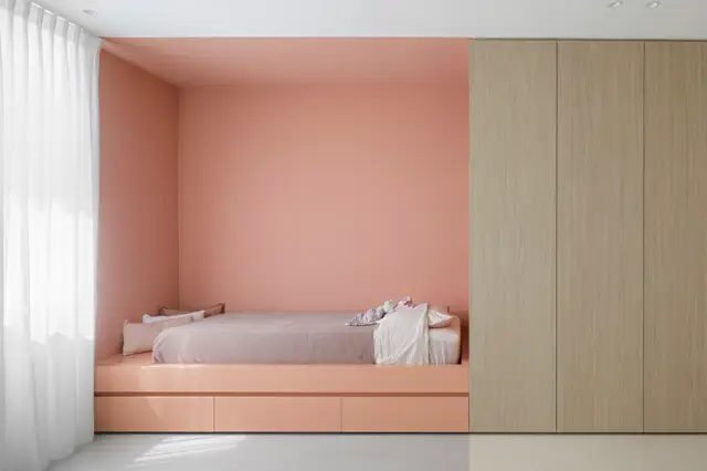 Een strakke slaapkamer met een ingebouwde bedniche in warm roze, afgewerkt met naadloze wanden en opbergruimte. Minimalistische inrichting met natuurlijk licht en houtaccenten.