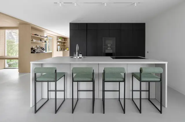 Strakke, lichte keukenruimte met een groot witte keukeneiland, naadloze lichte vloer en wand. Zwarte inbouwapparatuur, naturel houten kasten en groene designstoelen creëren een verfijnd contrast.