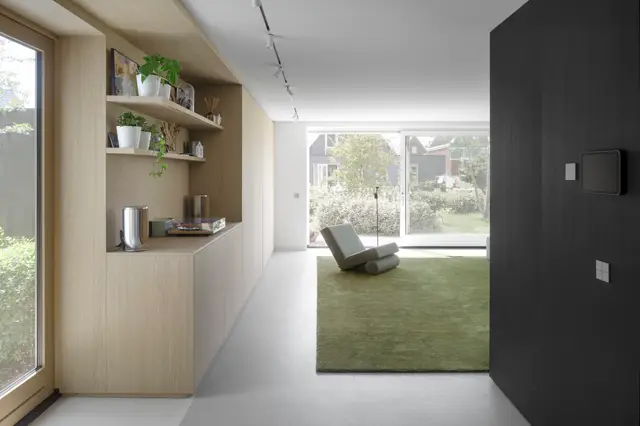 Een strak, licht wooninterieur met natuurlijke materialen, ingericht houten kasten met groene accenten en een grote raamwand die uitkijkt op het groen. De naadloze vloer loopt naadloos door naar buiten, creëerend een serene overgang.