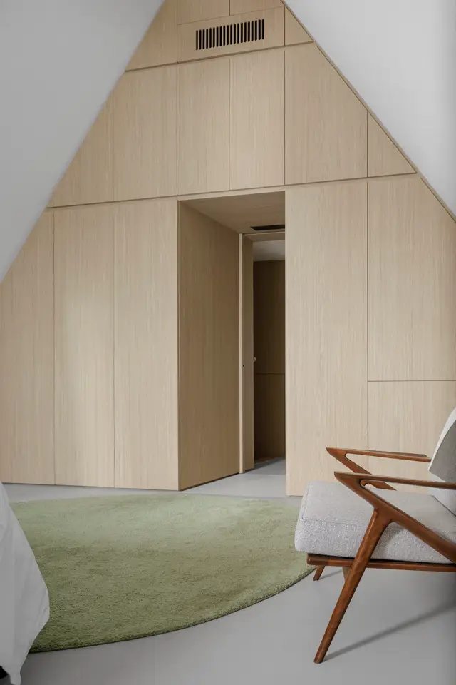 Een strak ontworpen ruimte met een grote houten wand in lichte houtnerf, naadloze afwerking en subtiele lijnen. Een groen tapijt en mid-century meubels creëren warmte in deze serene, verzorgde omgeving.
