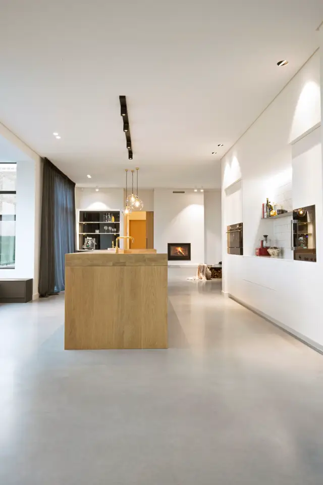 Ruime, licht interieur met een open keukenruimte. Een houten eiland staat centraal op een licht grijze naadloze vloer. De witte wanden en plafonds creëren een minimalistisch, schoon geheel met ingebouwde verlichting en natuurlijk daglicht.