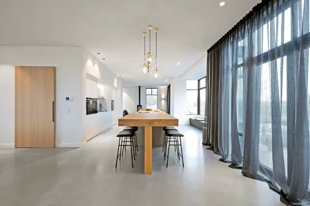 Ruim, minimalistisch interieur met naadloze lichte vloer die doorloopt van keuken naar woongebied. Grote raampartijen brengen natuurlijk licht naar binnen, elegante gouden hanglampen en strakke witte wanden creëren een serene luxe sfeer.