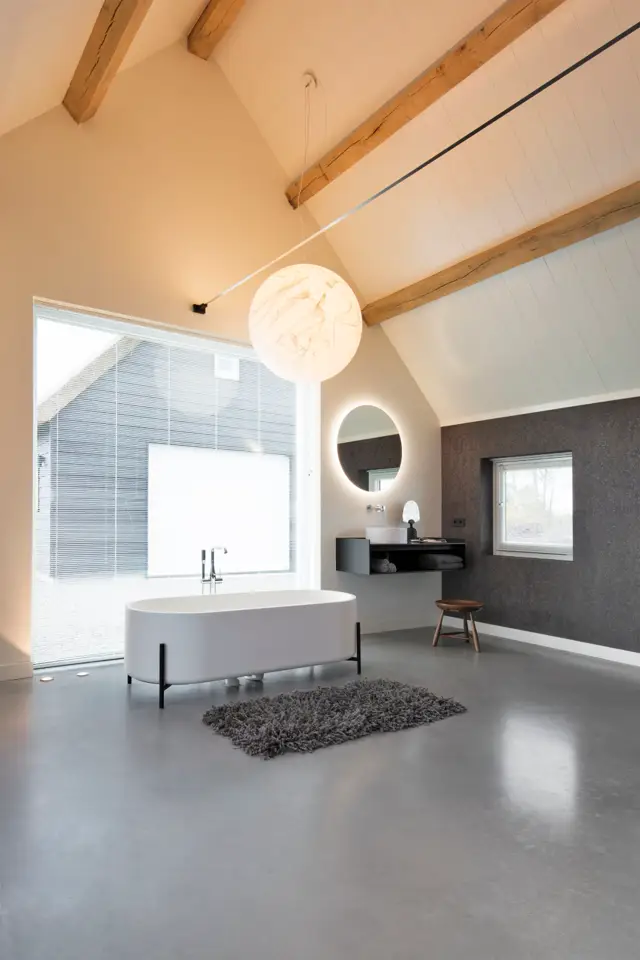 Ruime, lichte badkamer met een freestanding wit bad, naadloze grijze vloer, houten dakbalken en grote ramen. Minimalistische inrichting met donkergrijze accentwand en moderne verlichting.