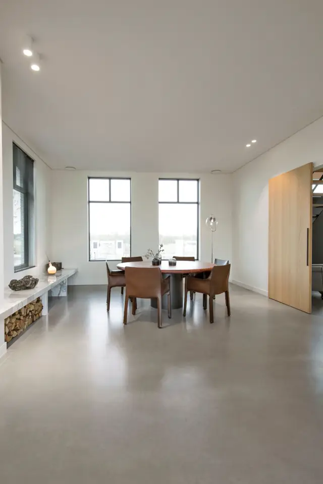 Ruime, heldere eetkamer met grote ramen, minimalistisch ingericht met houten tafel en bruine stoelen. De naadloze vloer loopt ononderbroken door de hele ruimte en geeft een zeer schoon, opgeruimd gevoel.