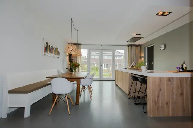 Lichte, strakke keuken-eetkamer met donkere naadloze vloer, witte wanden en houten accenten. Grote raampartij biedt veel daglicht en uitzicht op de tuin.