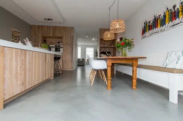 Strakke, lichte open ruimte met naadloze grijze vloer die naadloos overgaat van keuken naar eetgedeelte. Heldere belichting, minimalistische wandafwerking en georganiseerde interieurintegratie.