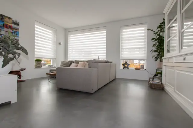 Ruime, licht ingerichte woonkamer met grote ramen en grote horizontale jaloezieën. Een grijze hoekbank staat centraal op een donkergrijze naadloze vloer die volmaakt glad en strak afloopt.