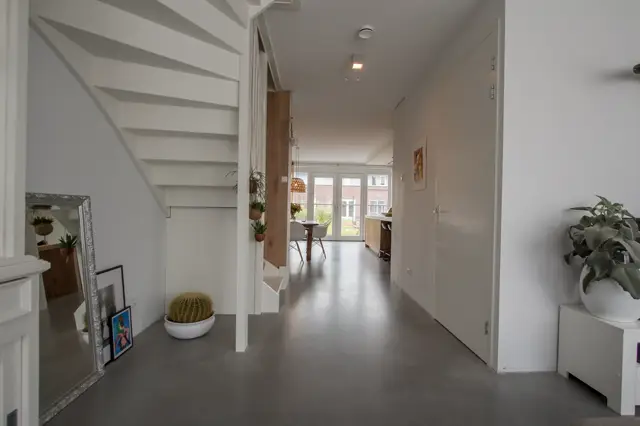 Een lichte, minimalistisch ingerichte hal met naadloze vloer in donkergrijs, witte wanden en scherpe architectonische lijnen. De ruimte ademt rust en opgeruimdheid uit met subtle groenaccenten.