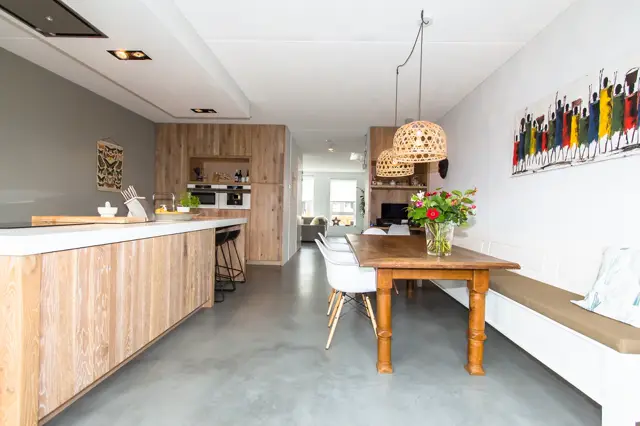Ruime, lichte open ruimte met naadloze grijze vloer die keuken en eethoek verbindt. Natuurlijke houttinten en minimalistisch design creëren een luxe, georganiseerde sfeer.