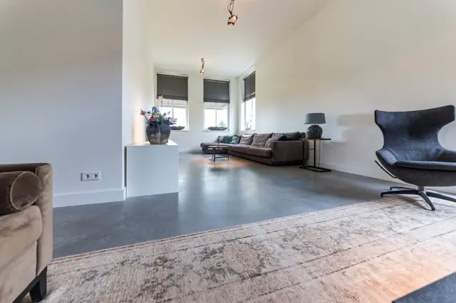Ruime, lichte woonkamer met een strakke naadloze vloer in donkergrijs. De ruimte straalt minimalistische elegantie uit met witte wanden, grote ramen en subtiele designmeubels.