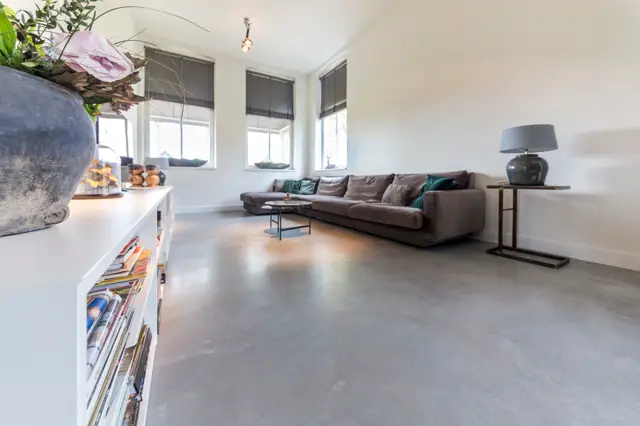 Ruime, lichte woonkamer met een brede naadloze vloer in warm grijs. Minimalistische inrichting met donkergrijs loungesofa, witte opbergkast en grote ramen die veel daglicht binnenbrengen. Alles ademt rust en ordening uit.