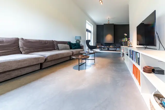 Ruime, licht ingevallen woonkamer met een strakke naadloze vloer in warm grijs. De ruimte ademt minimalisme en comfort met hoogwaardige afwerking.