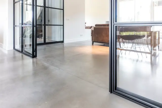 Ruime, lichte ruimte met glanzende naadloze vloer in warm grijs. Grote raampartijen met stalen frames zorgen voor heldere daglicht en uitzicht op terras. Minimaal ingericht met een lederen fauteuil.