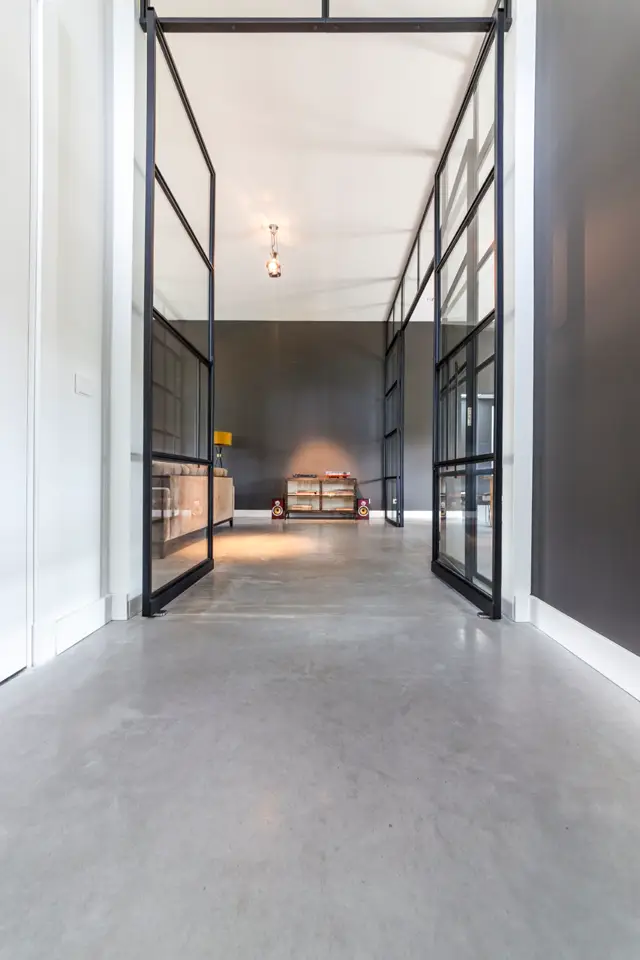 Een strak, licht interieur met grote geopende deuren in zwart staal. De naadloze vloer loopt glad door naar een open ruimte met subtiel warm licht en enkele designobjecten.